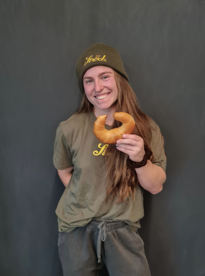 Eine lächelnde Person mit grüner Mütze und passendem Hemd hält ein halbmondförmiges Gebäck vor einem dunkelgrauen Hintergrund.