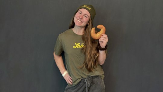 Eine lächelnde Person mit langen Haaren, die eine grüne Mütze und ein T-Shirt trägt, hält ein Hörnchen in einer Hand und steht vor einem schlichten dunkelgrauen Hintergrund.