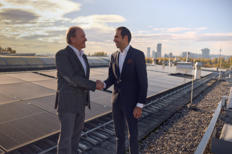 Zwei Geschäftsleute in Anzügen schütteln sich auf einem mit Solarzellen bedeckten Dach die Hände, während im Hintergrund bei teilweise bewölktem Himmel die Skyline der Stadt und Bäume zu sehen sind.