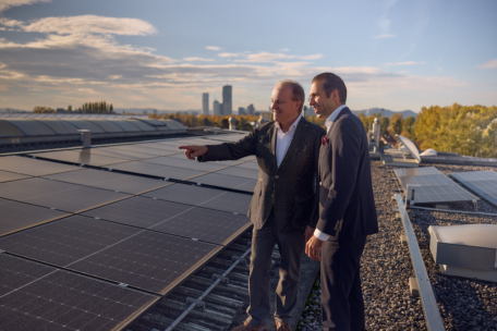 Zwei Männer in Anzügen stehen auf einem mit Solarzellen bedeckten Dach. Ein Mann zeigt in die Ferne, während der andere zusieht. Im Hintergrund ist die Skyline einer Stadt bei teilweise bewölktem Himmel zu sehen.