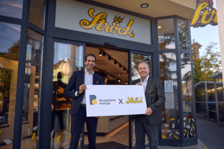 Zwei Männer in Anzügen stehen vor einer Bäckereifiliale und halten ein großes Schild mit der Aufschrift Burgenland Energie x Ströck. Über dem Eingang ist das Logo der Bäckerei zu sehen.