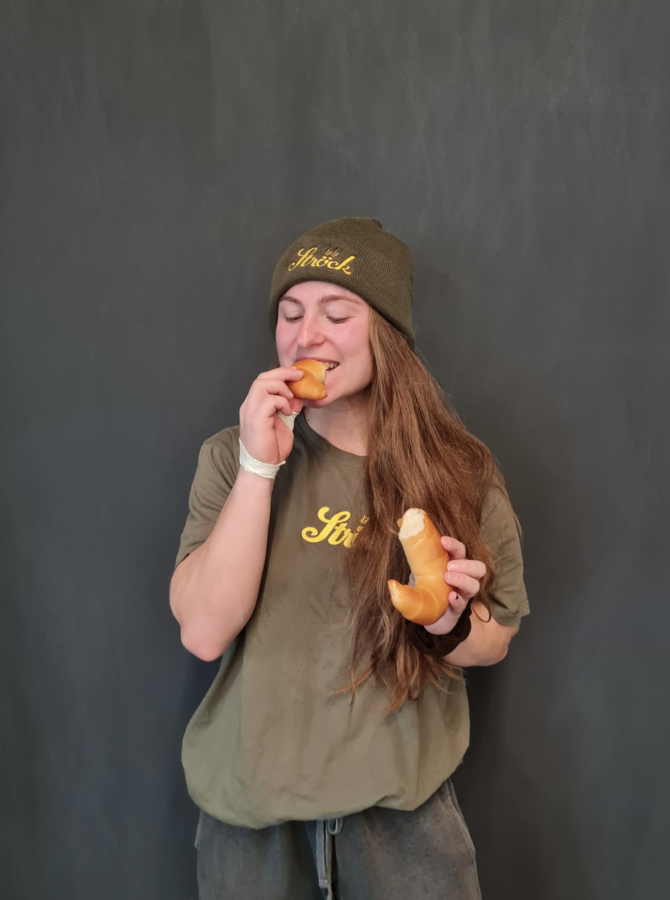 Eine Person mit langen braunen Haaren, die eine grüne Mütze und ein grünes T-Shirt trägt, lächelt mit geschlossenen Augen, während sie ein Croissant in der Hand hält und isst, während sie vor einem dunklen Hintergrund steht.