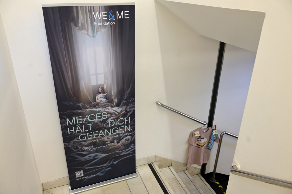 Ein vertikales Banner am oberen Ende einer Treppe zeigt eine Person, die auf einem unordentlichen Bett sitzt, mit einem deutschen Text, der lautet: ME/CFS hält dich gefangen. In der Nähe hängen T-Shirts am Treppengeländer. Die Wand ist weiß und schlicht.