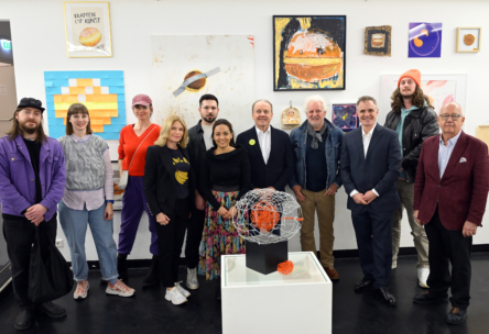 Eine Gruppe von elf Personen posiert in einer Kunstgalerie vor bunten, vielseitigen Kunstwerken, die an einer weißen Wand ausgestellt sind. Eine moderne Skulptur steht auf einem Sockel im Vordergrund.
