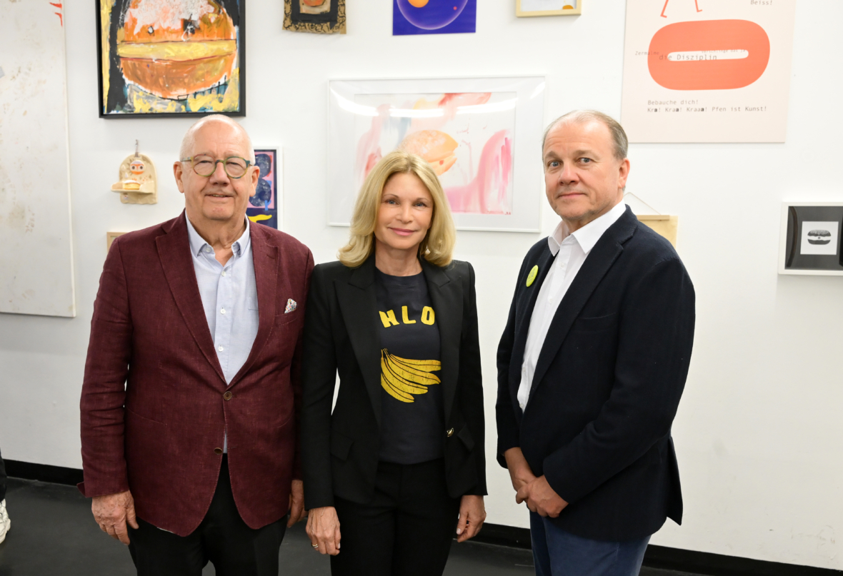 Drei Erwachsene stehen nebeneinander vor einer Wand mit bunter, vielseitiger Kunst. Der Mann links trägt einen kastanienbraunen Blazer, die Frau in der Mitte eine schwarze Jacke und ein Hemd mit Bananenmuster, und der Mann rechts trägt einen marineblauen Blazer.