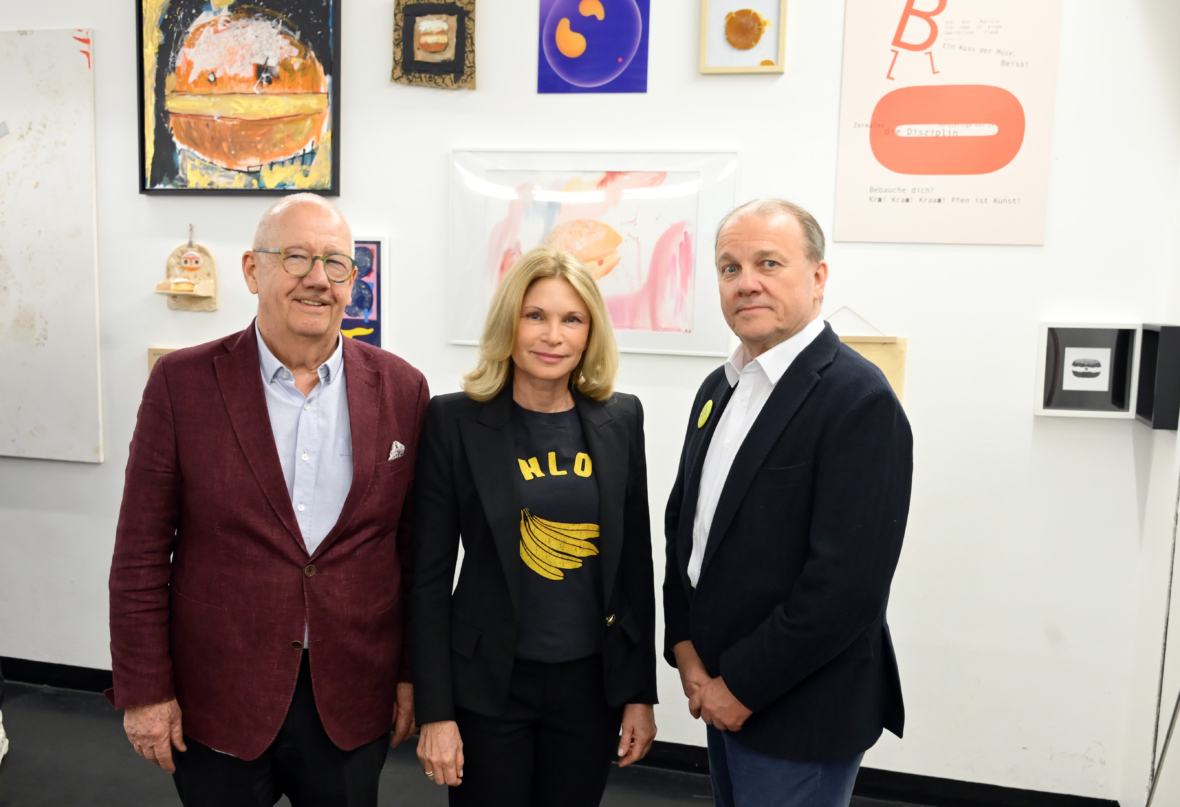 Drei Erwachsene stehen nebeneinander vor einer Wand, die mit bunten, abstrakten Kunstwerken bedeckt ist. Zwei Männer tragen Blazer; die Frau in der Mitte trägt eine schwarze Jacke und ein T-Shirt mit einer gelben Bananengrafik.