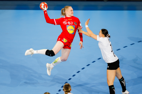 Eine Handballspielerin in einem roten Trikot springt, um den Ball zu schießen, während eine Verteidigerin in einem weiß-schwarzen Trikot ihren Arm zum Blocken hebt.