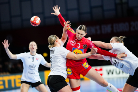 Eine Handballspielerin in einem roten Trikot springt, um den Ball zu werfen, während sie während eines intensiven Spiels von drei Gegnern in weißen Trikots verteidigt wird.
