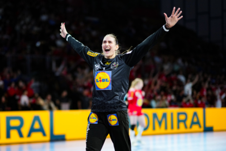Eine Handballspielerin in schwarzer Sportkleidung mit erhobenen Armen feiert auf dem Spielfeld. Auf ihrem Trikot ist das Lidl-Logo zu sehen. Im Hintergrund sind eine verschwommene Menschenmenge und eine weitere Spielerin in Rot zu sehen.