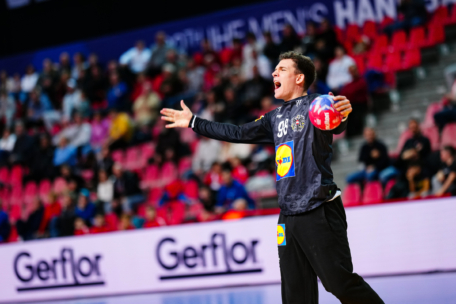 Ein Handballtorwart in einem schwarzen Trikot mit der Nummer 98 steht während eines Spiels mit ausgestreckten Armen und scheint zu schreien. Im Hintergrund sind Zuschauer auf roten Stadionsitzen zu sehen.