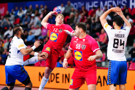 Ein Handballspieler in einem roten Trikot springt, um den Ball zu werfen, während er während eines Spiels von Gegnern in Weiß und Blau verteidigt wird. Im Hintergrund sind andere Spieler und Zuschauer zu sehen.