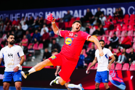 Ein Handballspieler in einem roten Trikot springt in der Luft und bereitet sich auf den Wurf vor, während Spieler in weißen Trikots zusehen. Im Hintergrund sind leere rote Sitze und ein verschwommenes Meisterschaftsbanner zu sehen.