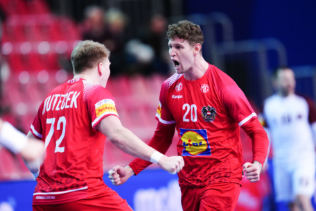 Zwei männliche Handballspieler in roten Trikots feiern leidenschaftlich auf dem Spielfeld, ballen die Fäuste und schreien, während im Hintergrund leere rote Sitze und ein weiterer Spieler verschwimmen.