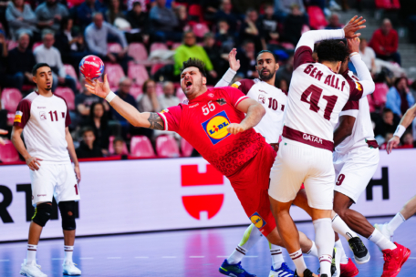 Ein Handballspieler in einem roten Trikot mit der Nummer 55 springt nach vorne und hält den Ball, während er von einem Spieler in einem weißen Trikot mit der Nummer 41 eng verteidigt wird. Andere Spieler und Zuschauer sind im Hintergrund zu sehen.