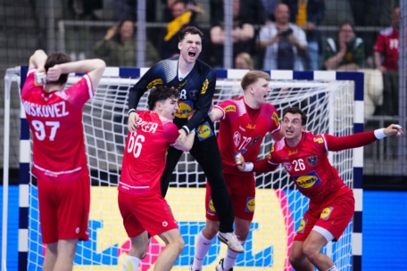 Vier Handballspieler in roten Trikots feiern freudig mit einem aufgeregten Torwart in Schwarz, der schreit und die Arme hochreißt, während sie sich in der Nähe des Torpfostens auf einem Hallenplatz versammeln. Im Hintergrund sind Zuschauer zu sehen.