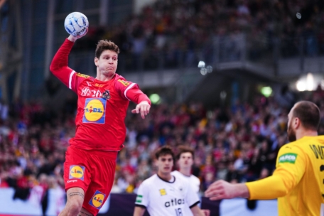 Ein männlicher Handballspieler in einem roten Trikot springt mit einem Ball in der erhobenen Hand durch die Luft und bereitet sich auf einen Schuss vor, während gegnerische Spieler in Weiß und ein Torwart in Gelb zusehen. Der Hintergrund zeigt eine unscharfe Menschenmenge in einer Halle.