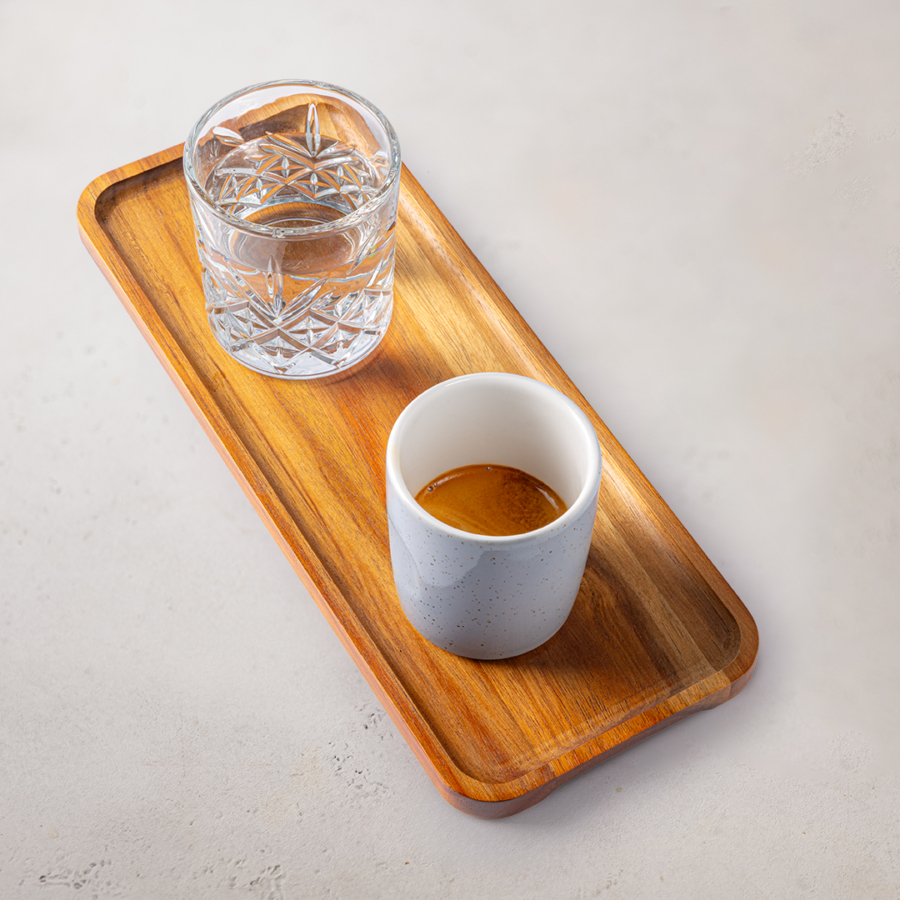 Eine kleine weiße Tasse Espresso und ein Glas Wasser stehen auf einem rechteckigen Holztablett, das auf einer hellen Fläche steht.