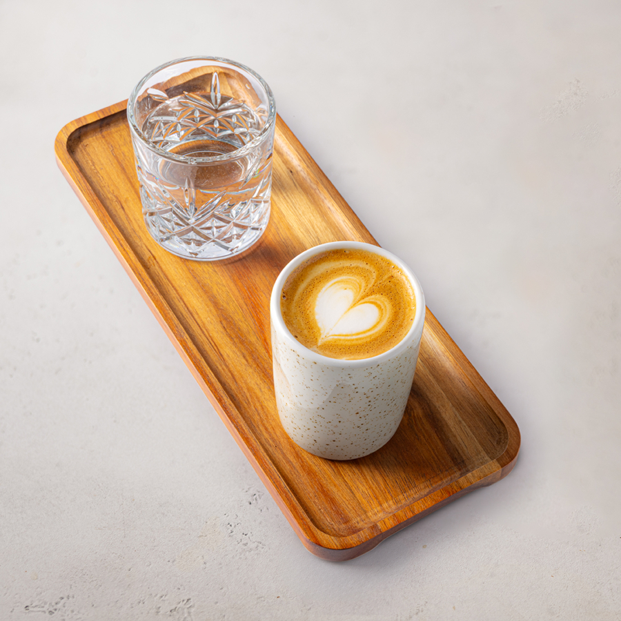 Ein Holztablett mit einem Glas Wasser und einer Keramiktasse mit einer herzförmigen Latte auf einer hellen Oberfläche.