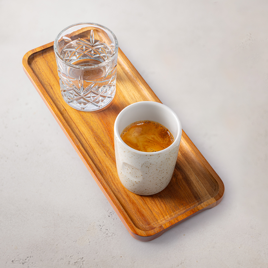 Eine kleine weiße Tasse Espresso und ein Glas Wasser stehen auf einem rechteckigen Holztablett, das auf einer hellen Fläche steht.