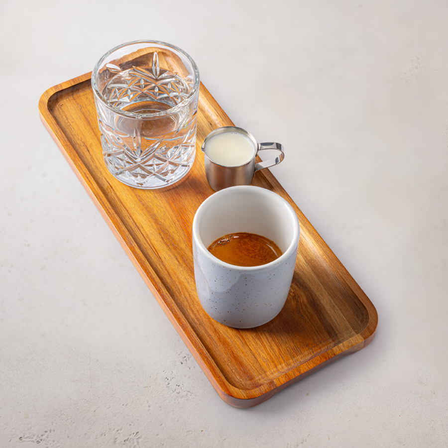 Auf einem Holztablett stehen ein Glas Wasser, ein Metallkännchen mit Milch und eine weiße Tasse mit Espresso, die ordentlich auf einer hellen Fläche angeordnet sind.