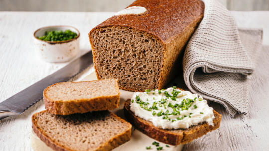 Ein Laib Schwarzbrot mit zwei aufgeschnittenen Scheiben, eine davon bestrichen mit Frischkäse und gehacktem Schnittlauch. Ein Messer, eine Schale mit Schnittlauch und eine gefaltete Stoffserviette liegen neben dem Brot auf einer hellen Fläche.