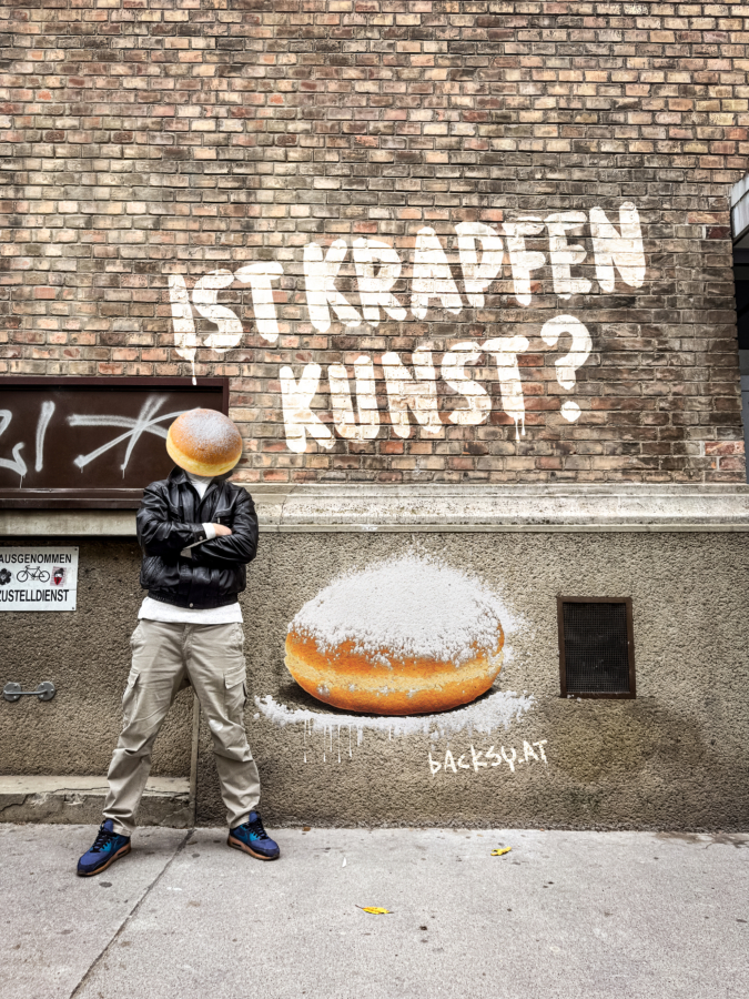 Eine Person steht an einer Backsteinmauer und hat ihren Kopf durch einen gepuderten Donut ersetzt. Auf dem Graffiti über ihr steht IST KRAPFEN KUNST? und ein großer Donut ist neben dem deutschen Text BACKSY.ART an die Wand gemalt.