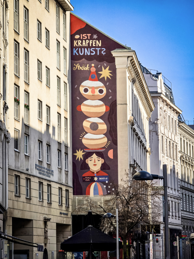 Ein hohes Wandbild an einem städtischen Gebäude zeigt einen skurrilen Stapel mit Gelee gefüllter Donuts über einer stilisierten Frau. Das Wandbild enthält den deutschen Text Ist Krapfen Kunst? und bunte, verspielte Motive. Andere Gebäude und ein klarer Himmel sind zu sehen.