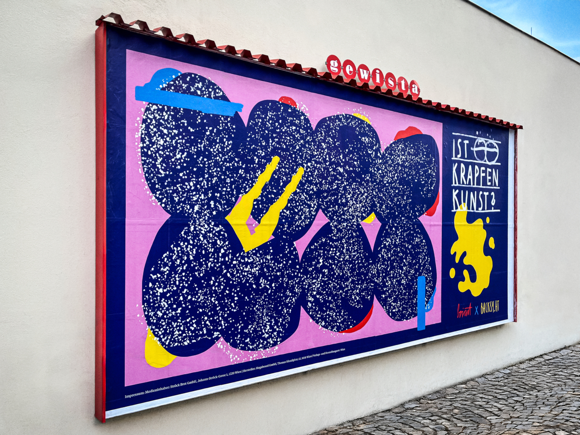 Eine farbenfrohe Plakatwand im Freien zeigt abstrakte blaue und gelbe Formen mit weißen Sprenkeln. Auf der rechten Seite steht "Ist Krapfen Kunst?", darunter befindet sich ein weiterer kleinerer Text mit kunstvollen gelben Sprenkeln.