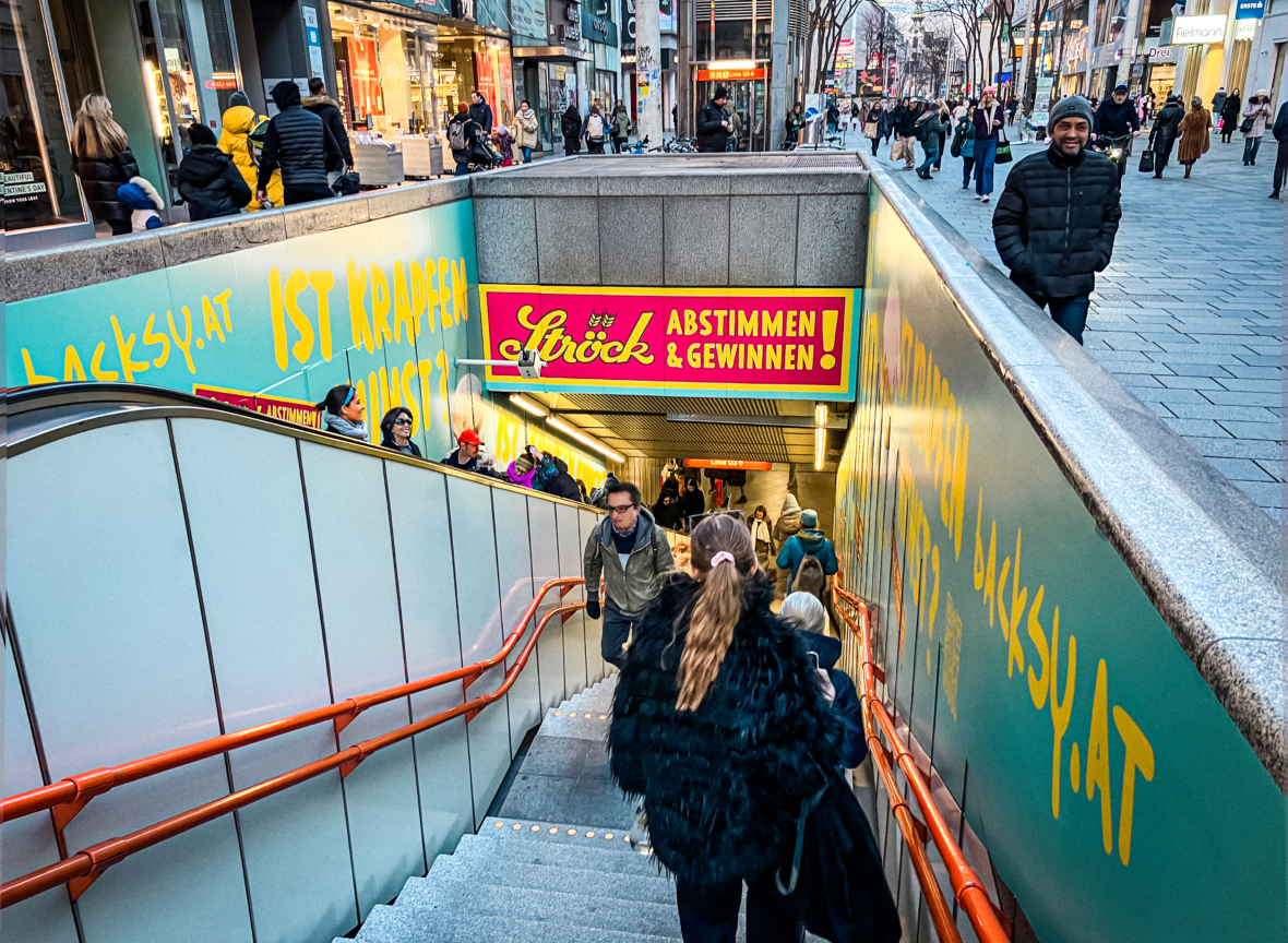 Menschen steigen eine belebte Außentreppe hinab und hinauf, die zu einem unterirdischen Bereich führt, umgeben von bunter Werbung in deutscher Sprache. Einkäufer und Fußgänger gehen eine mit Geschäften und hellen Schildern gesäumte Stadtstraße entlang.