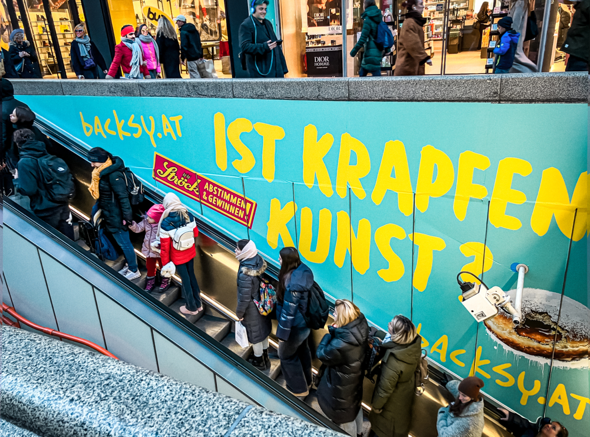 Menschen fahren auf einer Rolltreppe in einem Einkaufszentrum neben einer leuchtend blauen Wand mit der großen gelben deutschen Aufschrift IST KRAPFEN KUNST? und dem Bild eines teilweise in Kaffee getauchten Donuts.
