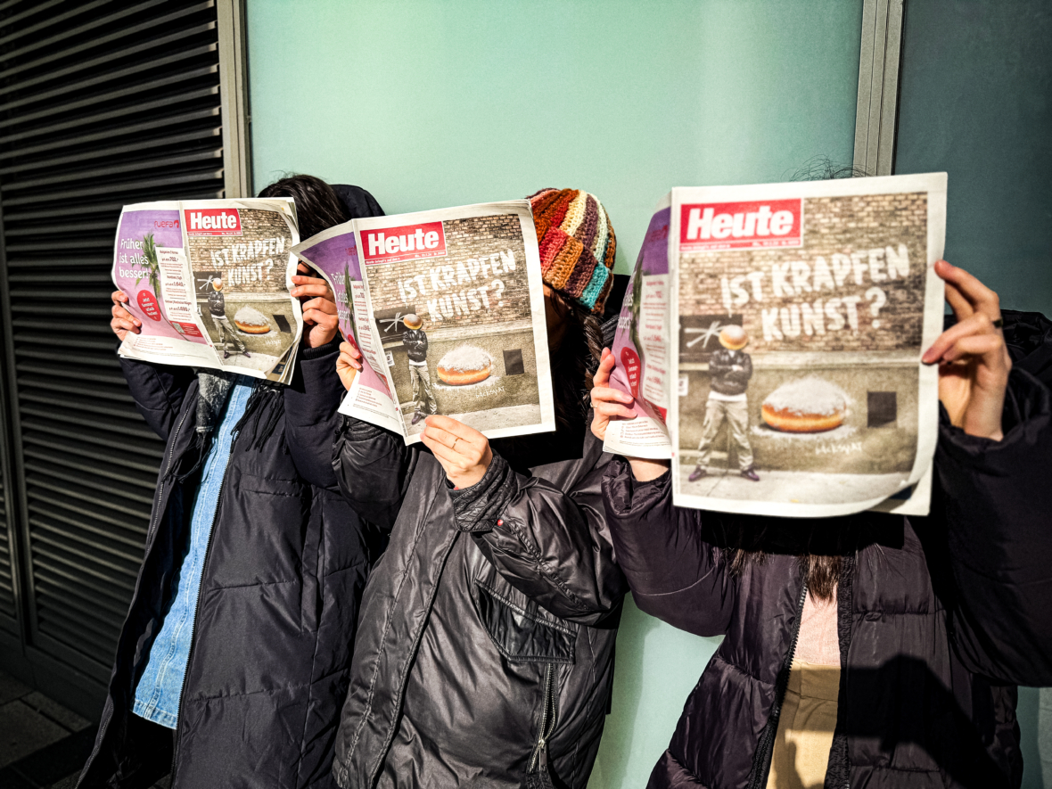 Drei Personen, die nebeneinander stehen, bedecken ihre Gesichter mit Zeitungen, auf deren Titelseite eine Mauer, Graffiti und ein großes Gebäck abgebildet sind. Die Schlagzeile lautet IST KRAPFEN KUNST? Sie tragen Winterjacken und Mützen.