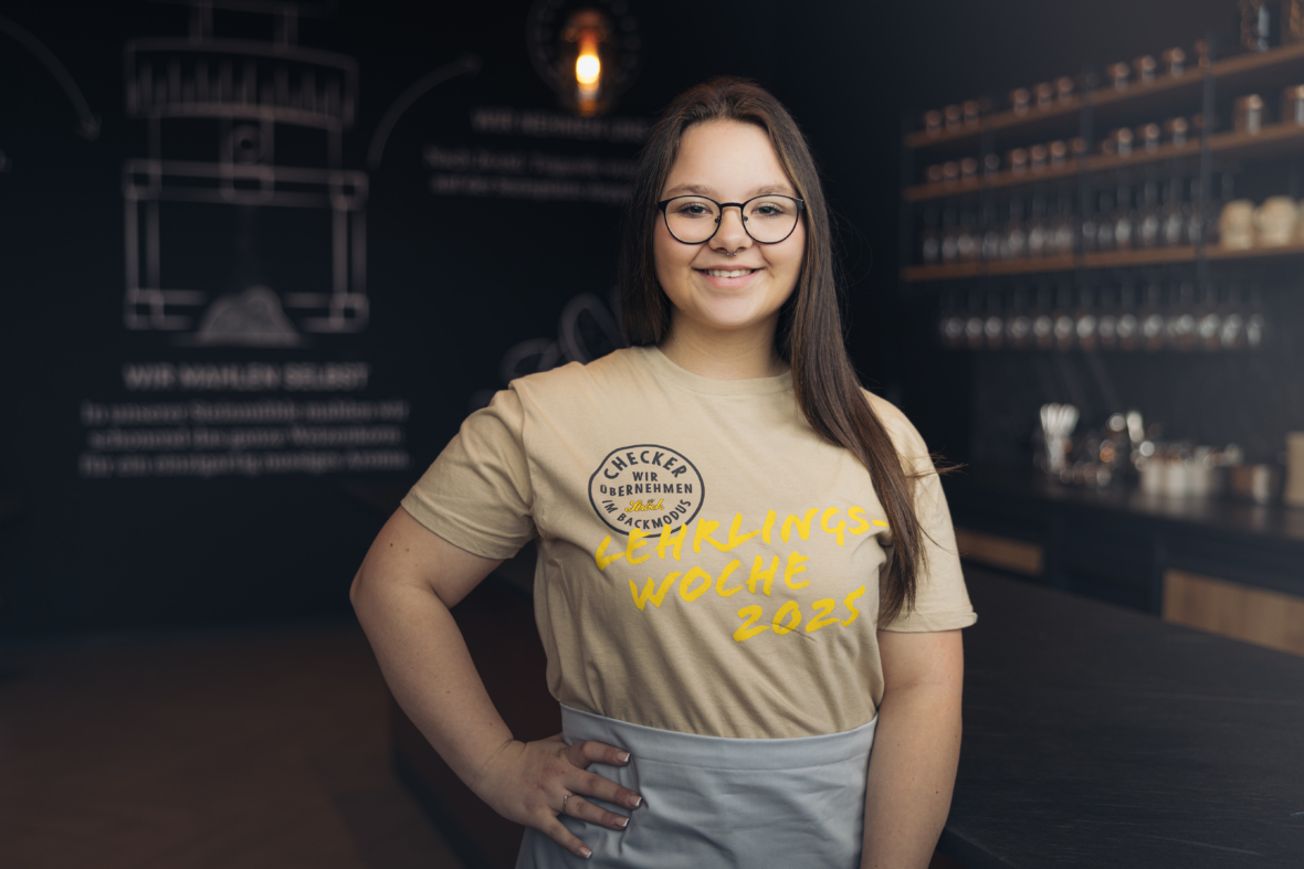 Eine junge Frau mit langen braunen Haaren und Brille steht lächelnd in einem modernen Cafe. Sie trägt ein beiges T-Shirt mit der Aufschrift "Lehrlings Woche 2025" und eine Schürze mit Regalen und einer Kreidetafel im Hintergrund.