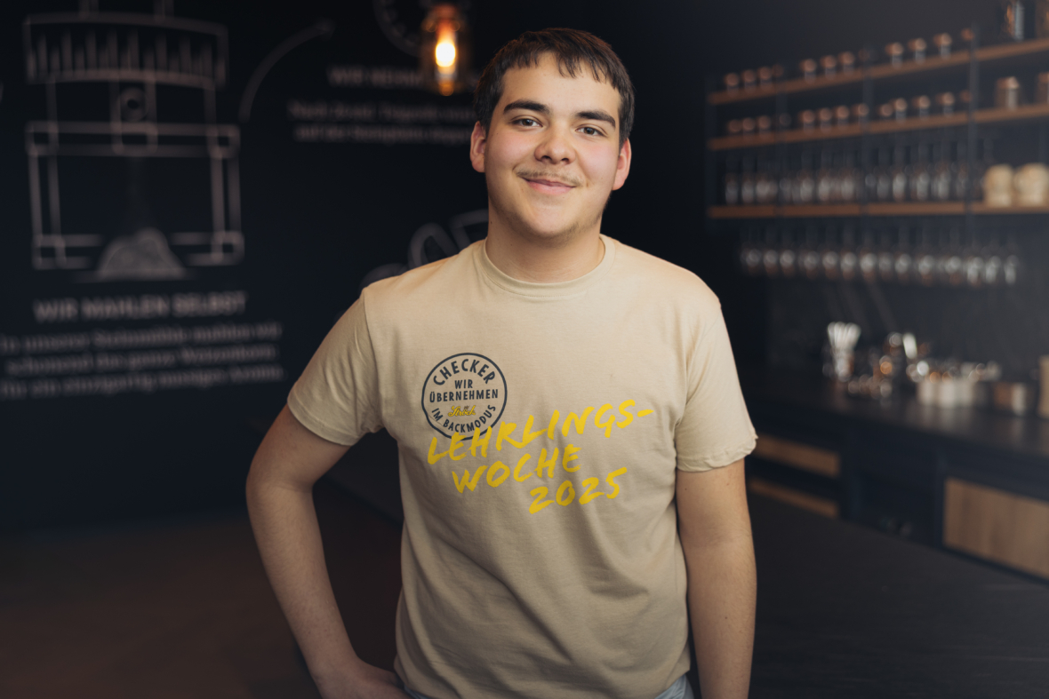 Ein lächelnder junger Mann steht in einer modernen Bar und trägt ein beiges T-Shirt mit der Aufschrift Lehrlings-Woche 2025 und Gereckte Übernahmen. Im Hintergrund sind Regale mit Gläsern und Flaschen zu sehen.