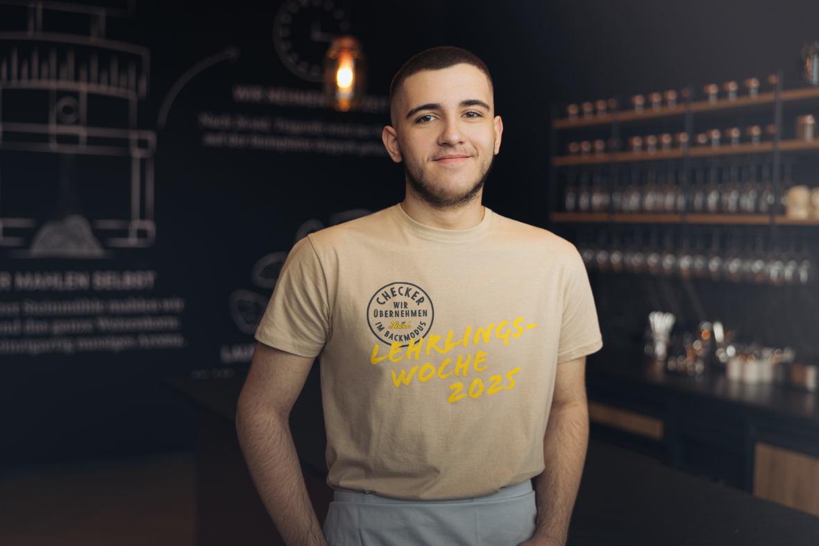 Ein junger Mann steht in einem Cafe und lächelt in die Kamera. Er trägt ein beiges T-Shirt mit dem gelben Aufdruck LEHRLINGS-WOCHE 2023, im Hintergrund sind Regale mit Tassen und Gläsern zu sehen.
