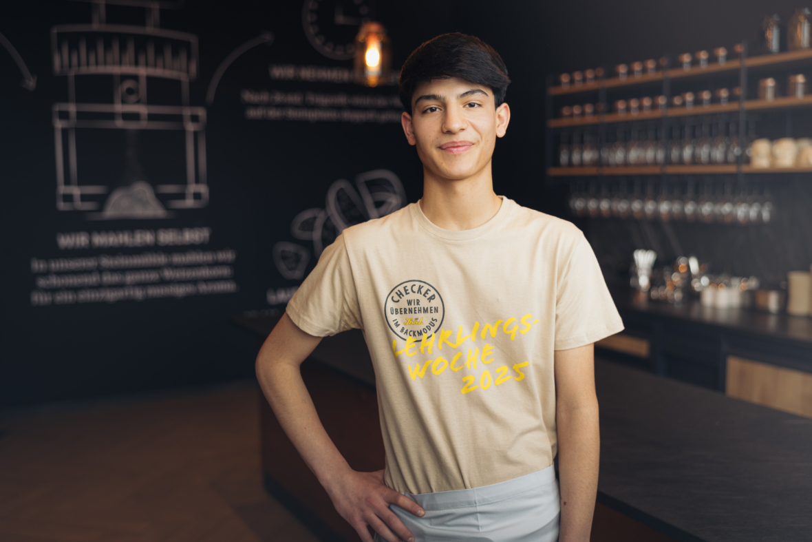 Eine junge Person mit kurzen dunklen Haaren steht selbstbewusst in einem modernen Café und trägt ein beiges T-Shirt mit der gelben Aufschrift "Lehrlingswoche 2023". Im Hintergrund befinden sich Regale mit Tassen und eine Kreidetafelwand.