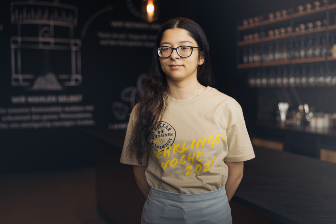 Eine junge Frau mit langen dunklen Haaren und Brille steht in einem modernen, schwach beleuchteten Café. Sie trägt ein beiges T-Shirt mit gelber Schrift und eine hellgraue Schürze, im Hintergrund stehen Regale und eine Kreidetafel.
