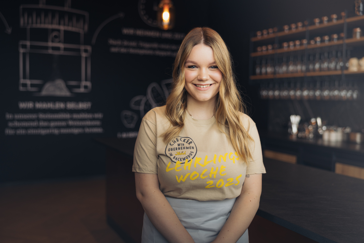 Eine junge Frau mit langen blonden Haaren steht lächelnd in einem modernen Café. Sie trägt ein beiges "Lehrlings Woche 2023"-T-Shirt und eine weiße Schürze. Hinter ihr stehen Regale mit Tassen und Wandmalereien im Stil einer Kreidetafel.