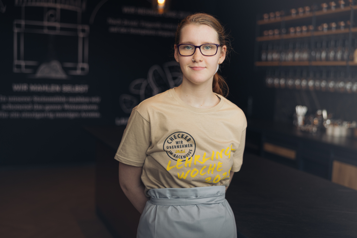 Eine junge Frau mit Brille und Pferdeschwanz steht in einem Innenraum und trägt ein beiges T-Shirt mit dem Aufdruck Lehrlings Woche 2021. Der Hintergrund ist ein modernes, schwach beleuchtetes Café oder Geschäft mit Regalen und Utensilien.