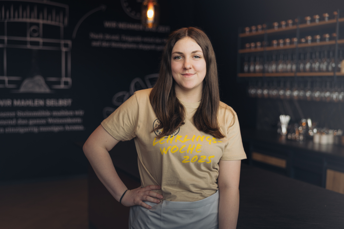 Eine junge Frau mit langen braunen Haaren steht selbstbewusst in einem modernen Café, trägt ein beiges Hemd mit der Aufschrift Lehrlingswoche 2023 und eine weiße Schürze, die Hand in der Hüfte und ein leichtes Lächeln.