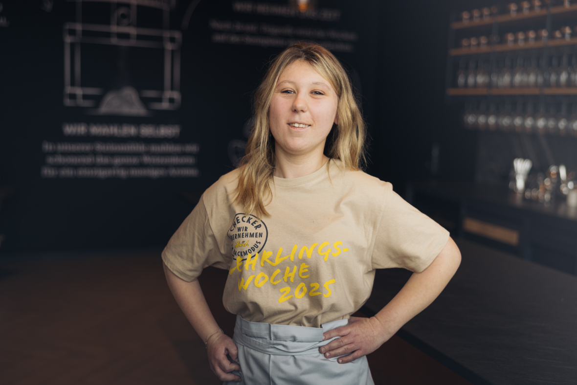 Eine Frau mit langen blonden Haaren steht in einem Haus und stützt lächelnd die Hände in die Hüften. Sie trägt ein beiges T-Shirt mit der Aufschrift Lieblingswoche 2025 und eine helle Schürze. Im Hintergrund ist ein schummriges Café oder Restaurant zu sehen.