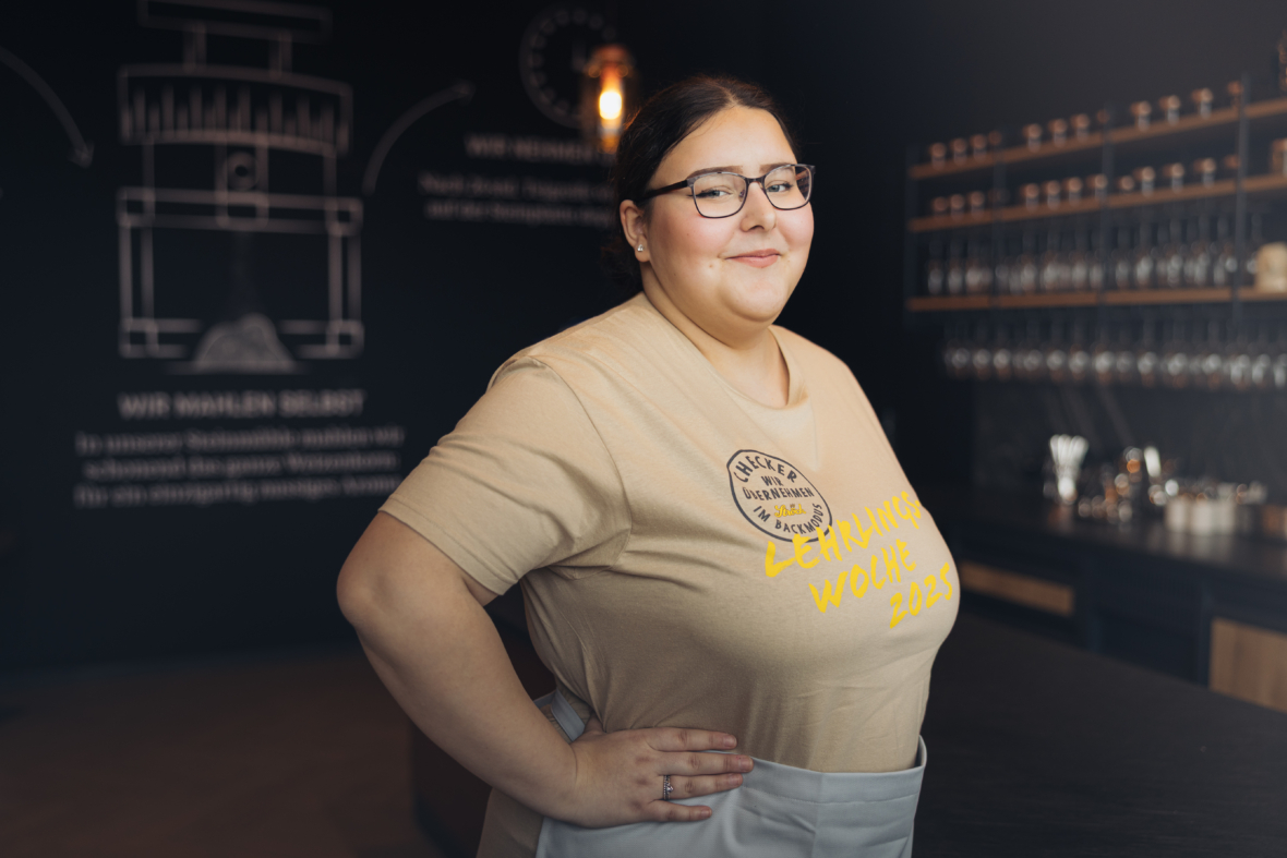 Eine Frau mit Brille und zurückgebundenen dunklen Haaren lächelt selbstbewusst, als sie hinter dem Tresen eines modernen Cafés steht. Sie trägt ein beiges T-Shirt mit einem Logo und gelber Schrift sowie eine hellgraue Schürze. Hinter ihr stehen Regale an der Wand.