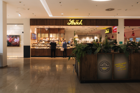 Zwei Personen stehen an der Theke einer Bäckerei namens "Ströck" in einem Einkaufszentrum. Die gut beleuchtete Bäckerei bietet Gebäck und Brot an, im Vordergrund stehen Pflanzen und eine Holzschranke.