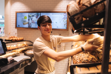 Ein junger Mann mit Handschuhen und einem beigen T-Shirt lächelt, während er in einer Bäckerei Brote auf einem Regal mit verschiedenen Backwaren und einer digitalen Speisekarte im Hintergrund anordnet.