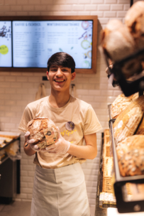 Ein junger Bäckereiarbeiter in Schürze und Handschuhen lächelt, während er einen Laib Brot in der Hand hält. Er steht vor Regalen, die mit verschiedenen Brotsorten gefüllt sind, und im Hintergrund ist eine Tafel mit der Speisekarte zu sehen.