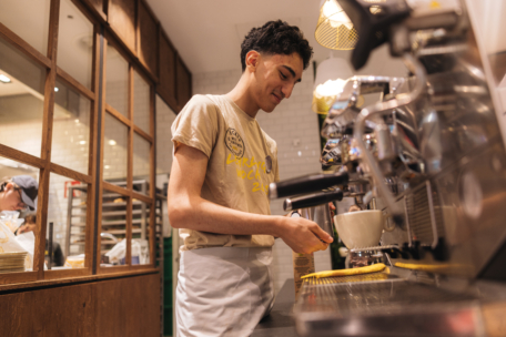 Ein junger Barista in T-Shirt und Schürze lächelt, während er hinter einem Tresen Kaffee zubereitet und eine Espressomaschine in einem gemütlichen Café mit Holz- und Kacheldekor bedient.