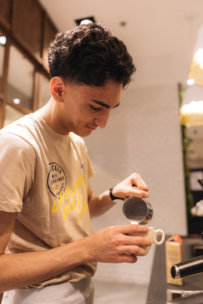 Ein junger Mann mit kurzen lockigen Haaren gießt gedämpfte Milch aus einem Metallkrug in eine Tasse und kocht Kaffee in einer modernen Küche mit weißen Fliesen und Holzakzenten.