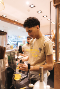Ein junger Barista in einem beigen T-Shirt bereitet hinter einem Tresen in einem gut beleuchteten Café ein Kaffeegetränk zu, während im Hintergrund ein weiterer Mitarbeiter und Backwaren zu sehen sind.