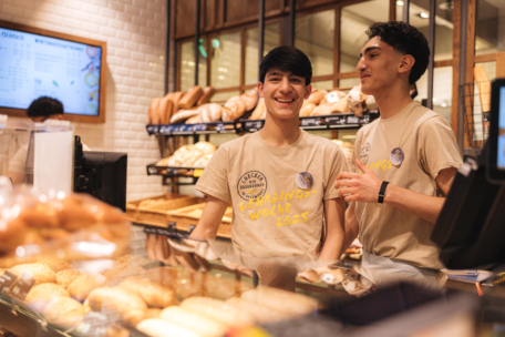 Zwei junge Männer in Bäckeruniformen lächeln und unterhalten sich hinter dem Tresen einer Bäckerei, mit Regalen voller Brot und Gebäck im Hintergrund.