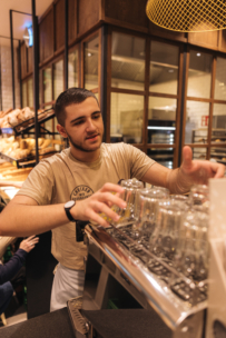 Ein junger Mann in einem beigen T-Shirt stellt saubere Gläser auf eine Espressomaschine in einem gemütlichen, modernen Café, während im Hintergrund Brot in Regalen steht.