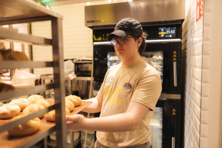 Eine Person mit Brille, Mütze und beigefarbenem T-Shirt stellt Tabletts mit frisch gebackenen Brötchen auf ein Gestell in einer Bäckereiküche, wobei im Hintergrund Öfen und Geräte zu sehen sind.